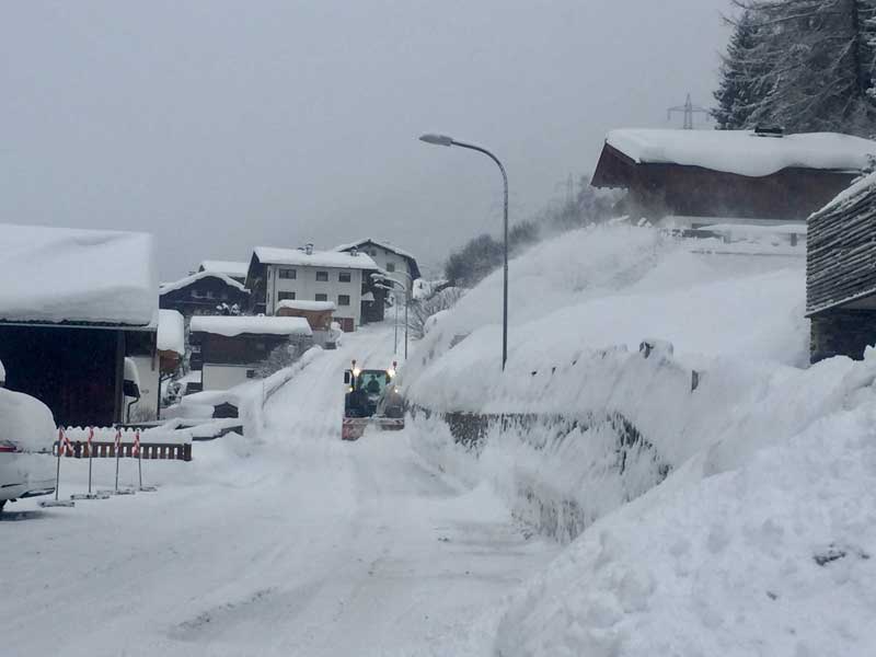 Schneepflug räumt Schnee in einem Bergdorf, hohe Schneewände links und rechts.