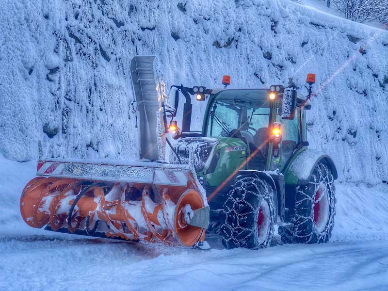 Schneefräse im Einsatz auf vereister Straße, Schnee reflektiert blaues Licht der Umgebung.