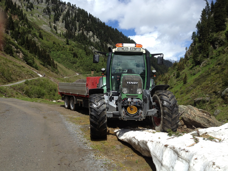 Fendt-Traktor mit Anhänger parkt auf einem alpinen Bergweg neben Schneeresten.